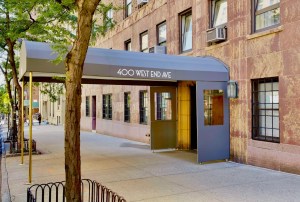 The sidewalk entrance to 400 West End Ave, featuring a large grey curved awning supported by gold poles. The building is a multi-story brownstone with dark-framed windows and air conditioning units. A small tree stands in the foreground on a clean concrete sidewalk.